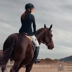 Cavalière en tenue d'équitation, casque noir, effectuant un exercice de dressage galop 7 sur un cheval brun. Arrière-plan : manège, bâtiments et ciel nuageux.