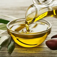 Un filet d'huile d'olive dorée coule d'une carafe dans un bol. Des olives et des feuilles d'olivier complètent la scène, évoquant la pureté de ce trésor végétal.