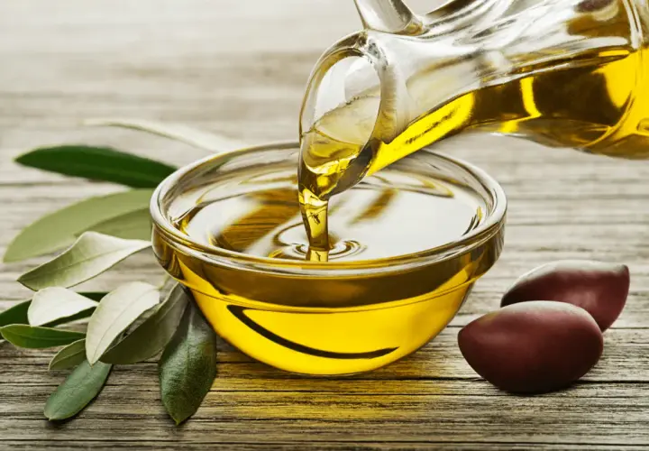 Un filet d'huile d'olive dorée coule d'une carafe dans un bol. Des olives et des feuilles d'olivier complètent la scène, évoquant la pureté de ce trésor végétal.