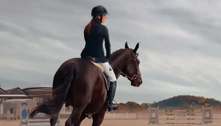Cavalière en tenue d'équitation, casque noir, effectuant un exercice de dressage galop 7 sur un cheval brun. Arrière-plan : manège, bâtiments et ciel nuageux.