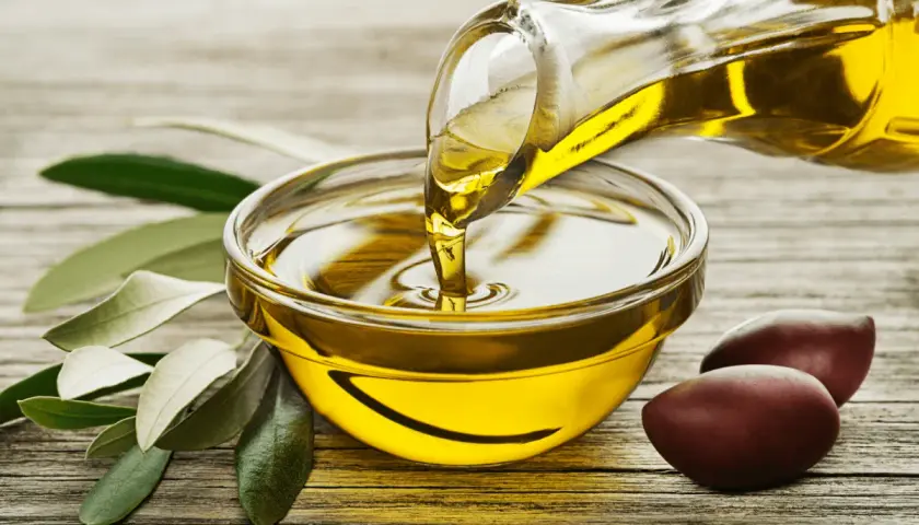 Un filet d'huile d'olive dorée coule d'une carafe dans un bol. Des olives et des feuilles d'olivier complètent la scène, évoquant la pureté de ce trésor végétal.