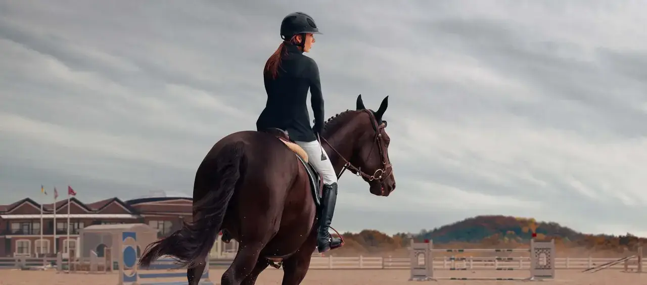 Cavalière en tenue d'équitation, casque noir, effectuant un exercice de dressage galop 7 sur un cheval brun. Arrière-plan : manège, bâtiments et ciel nuageux.