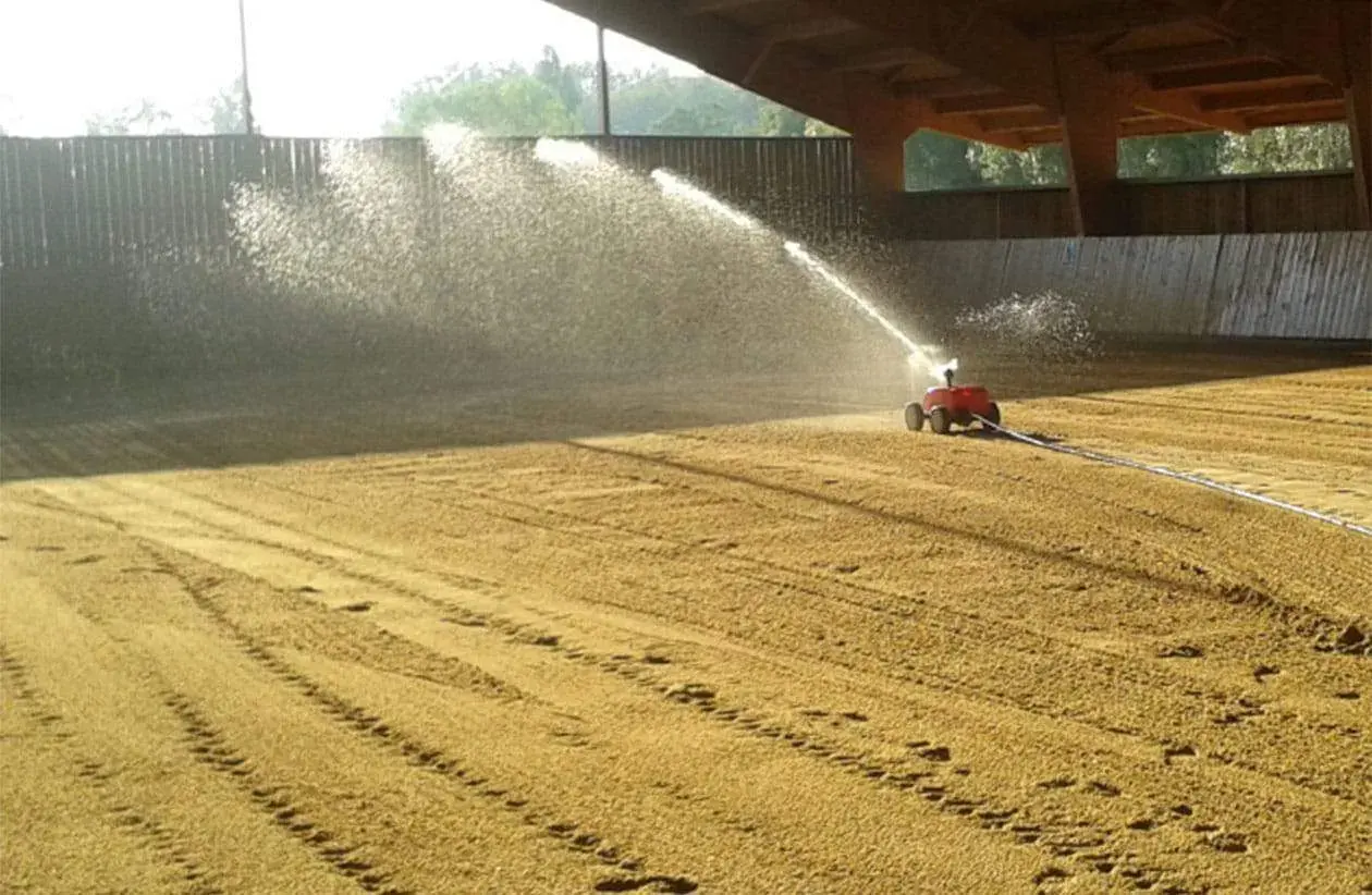 Arrosage d'une carrière équestre par un système mobile, créant un voile d'eau sur le sable.