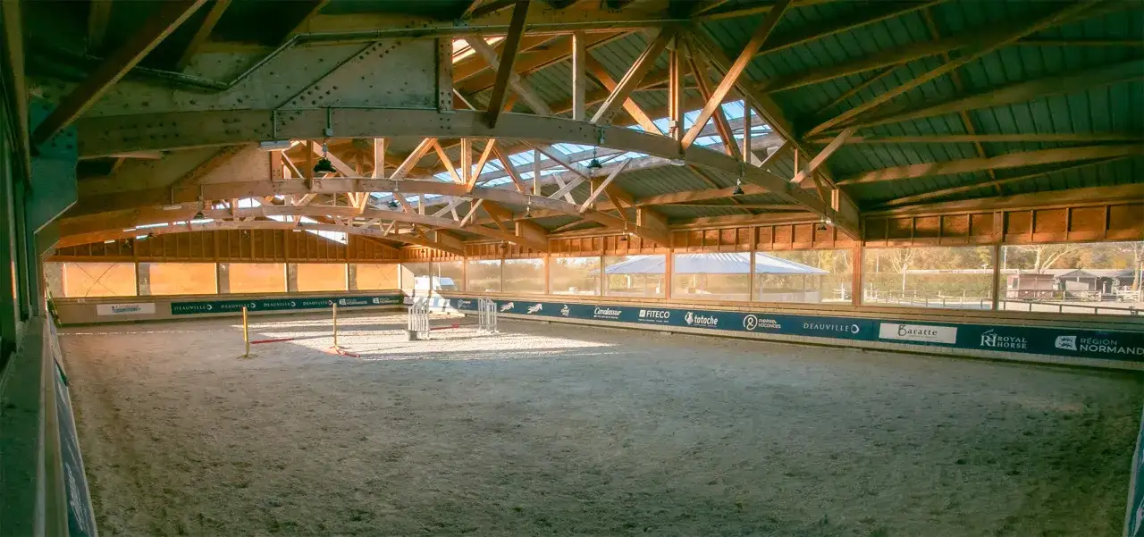 Int&eacute;rieur d'une carri&egrave;re de saut d'obstacle, avec une structure en bois et un toit vitr&eacute;. Le sable est visible au sol, pr&ecirc;t pour l'entra&icirc;nement.