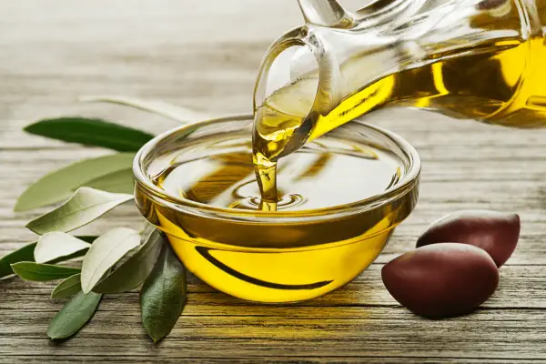 Un filet d'huile d'olive dorée coule d'une carafe dans un bol. Des olives et des feuilles d'olivier complètent la scène, évoquant la pureté de ce trésor végétal.